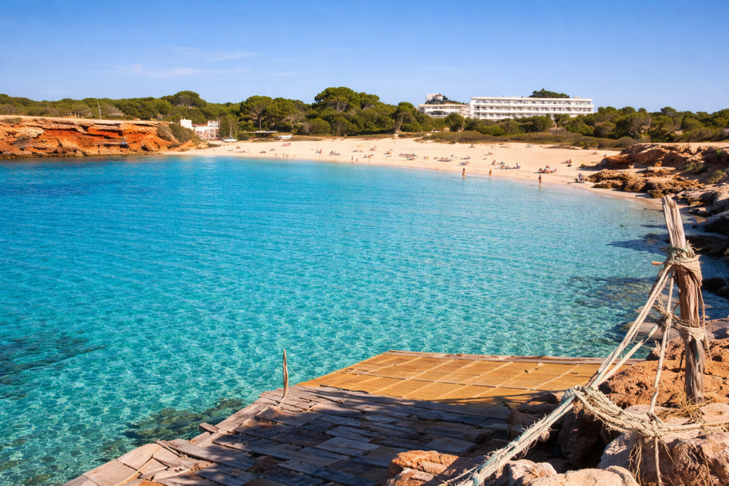 View of Cala Saona beach in Formentera with clear turquoise water, sandy shoreline, red rocky cliffs and a rustic wooden platform in the foreground under a bright blue sky.