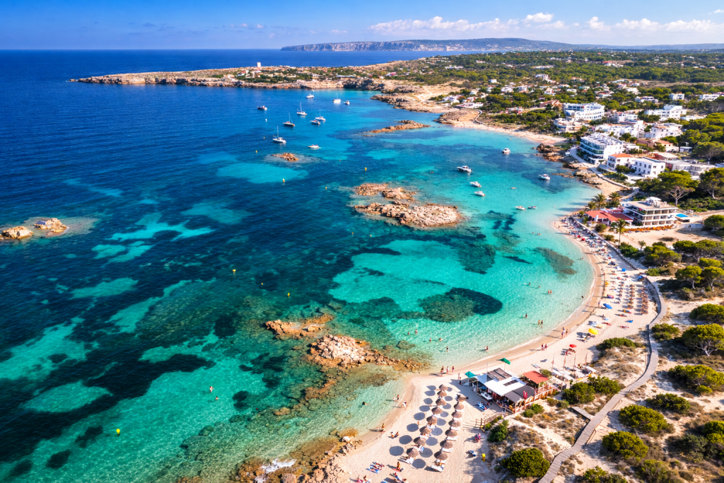 Aerial view of a Mediterranean resort coastline with turquoise water, rocky outcrops, sandy beach with sun loungers and umbrellas, and white seaside buildings along the shore.