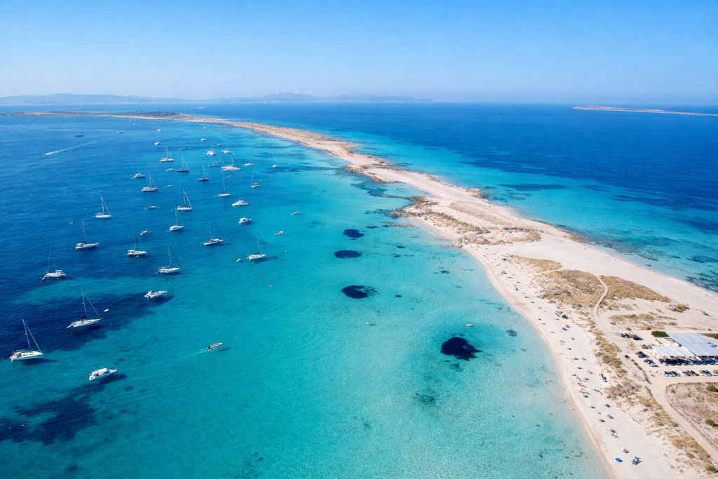 Aerial view of a narrow sandy peninsula surrounded by turquoise sea, with anchored yachts on one side and shallow crystal-clear water along the beach under a clear blue sky.
