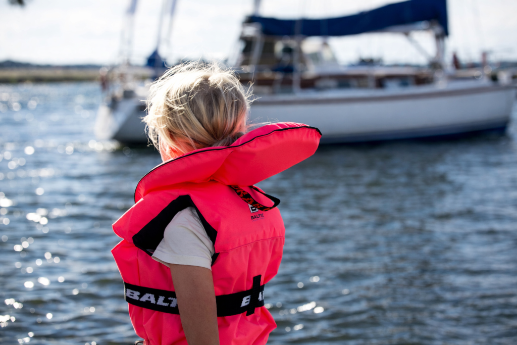 Un enfant portant un gilet de sauvetage rose vif se tient debout sur un bateau, regardant l'eau calme avec un voilier en arrière-plan par une journée ensoleillée.