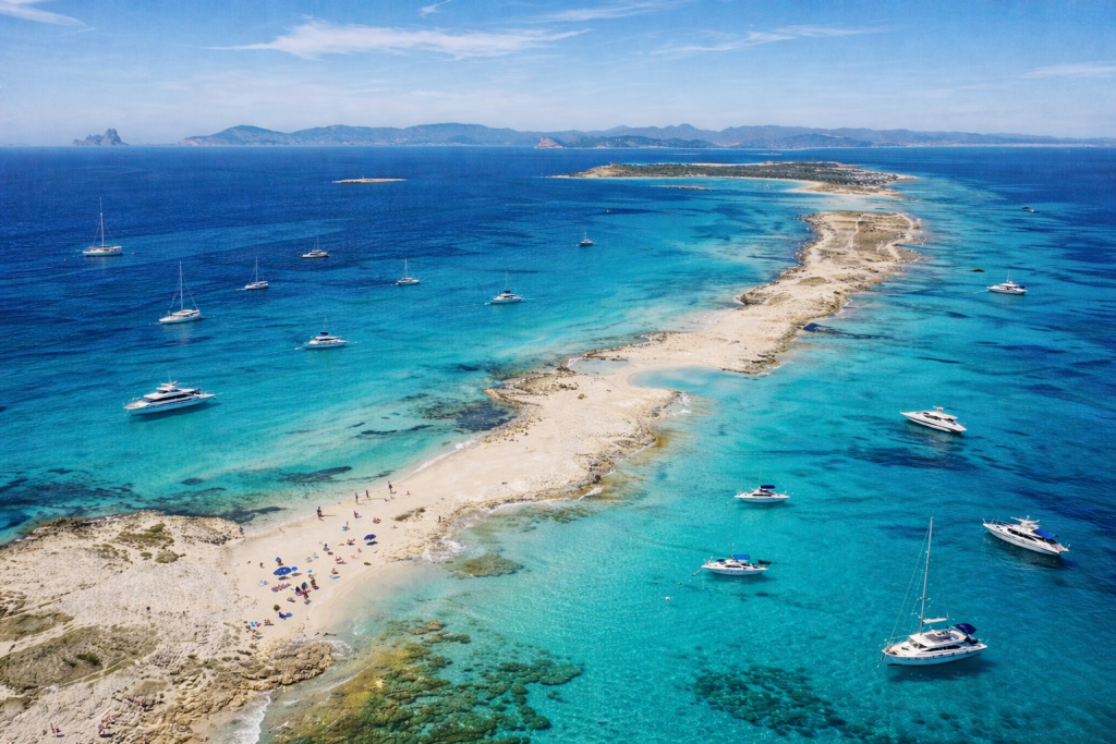 Aerial view of a narrow sandy island surrounded by crystal-clear turquoise water, with multiple yachts and boats anchored nearby and small groups of people relaxing along the shoreline.