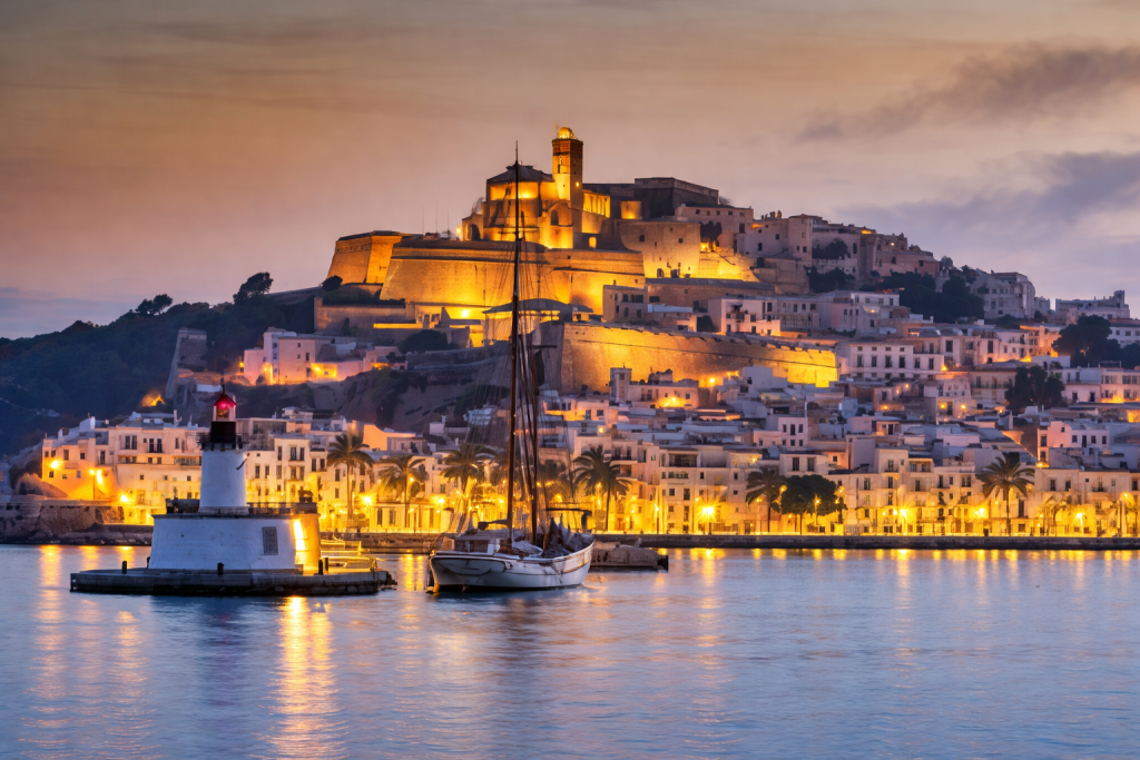 Abendlicher Blick auf eine mediterrane Küstenstadt mit beleuchteten historischen Gebäuden auf einem Hügel, einem Leuchtturm und einem Segelboot im Hafen sowie ruhigem Wasser, das im Sonnenuntergang warmes goldenes Licht reflektiert.