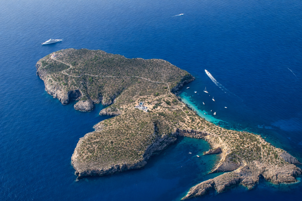 Luftaufnahme einer zerklüfteten Mittelmeerinsel mit felsigen Klippen, gewundenen Pfaden und spärlicher Vegetation, umgeben von tiefblauem Meer, auf dem Boote weiße Spuren im Wasser hinterlassen.