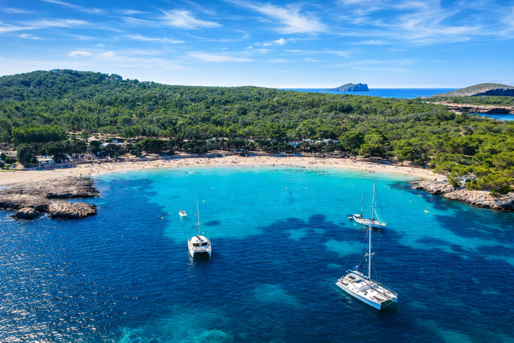 Luftaufnahme einer sichelförmigen Mittelmeerbucht mit klarem türkisfarbenem und tiefblauem Wasser, vor Anker liegenden Segelbooten, einem von Bäumen gesäumten Sandstrand und einer bewaldeten Küste unter strahlend blauem Himmel.