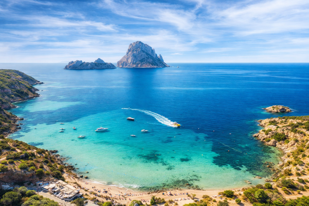 Luftaufnahme einer mediterranen Bucht mit klarem türkisfarbenem Wasser, Sandstrand und felsiger Küste, kleinen Booten, die in der Bucht vor Anker liegen, und einer großen Felseninsel, die sich in der Ferne unter strahlend blauem Himmel aus dem Meer erhebt.