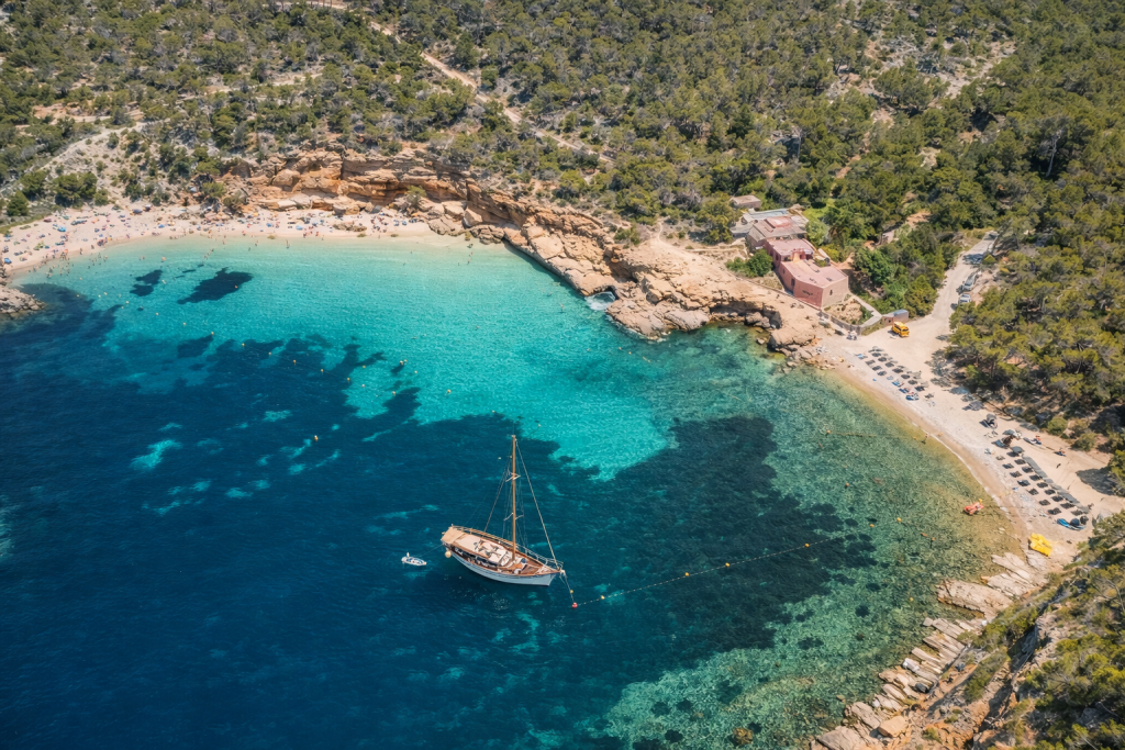 Luftaufnahme eines abgelegenen Mittelmeerstrandes mit klarem türkisfarbenem Wasser, felsiger Küste und einer kleinen Sandbucht, umgeben von dichten Pinienbäumen und einem Strandgebäude mit Sonnenliegen am Ufer.