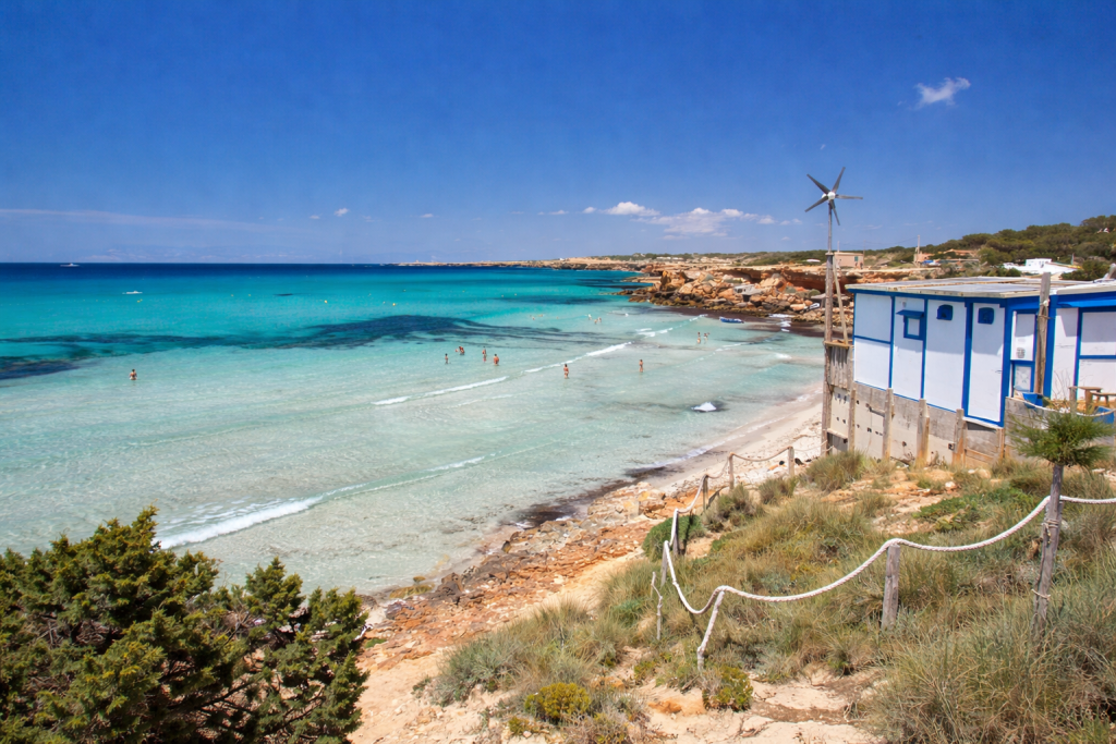 Ein mediterraner Küstenstrand mit klarem, türkisfarbenem Wasser und sanften Wellen, Menschen, die im seichten Wasser schwimmen, und eine weiß-blaue Strandhütte mit einer Windkraftanlage, die den Blick auf die sandige Küste und die felsige Küste freigibt.