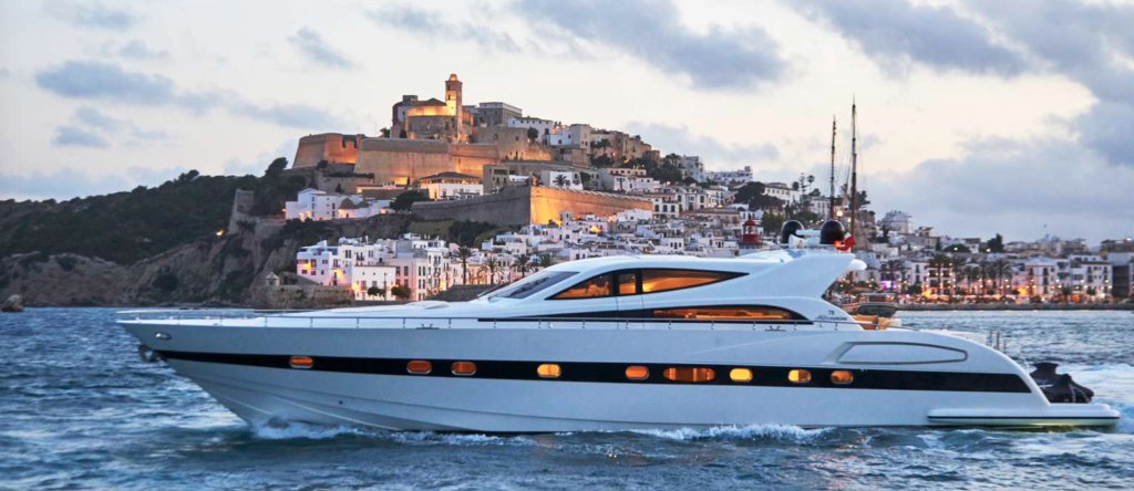Un lujoso yate a motor navegando en mar abierto con un pueblo costero y edificios históricos en la cima de una colina al fondo, con las luces reflejándose en el mar.