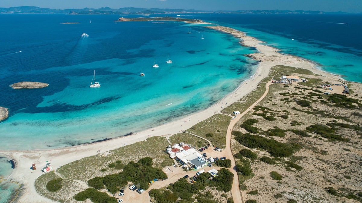 Aerial view of a narrow sandbar beach with clear turquoise water on both sides, boats anchored nearby, and coastal pathways with small buildings.
