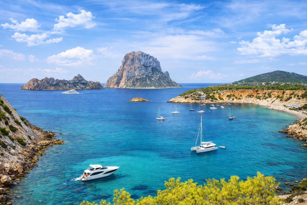 Mediterranean bay with turquoise water, sailboats and a motor yacht, surrounded by rocky coastline and green hills, with a large rocky island rising in the distance under a bright blue sky.