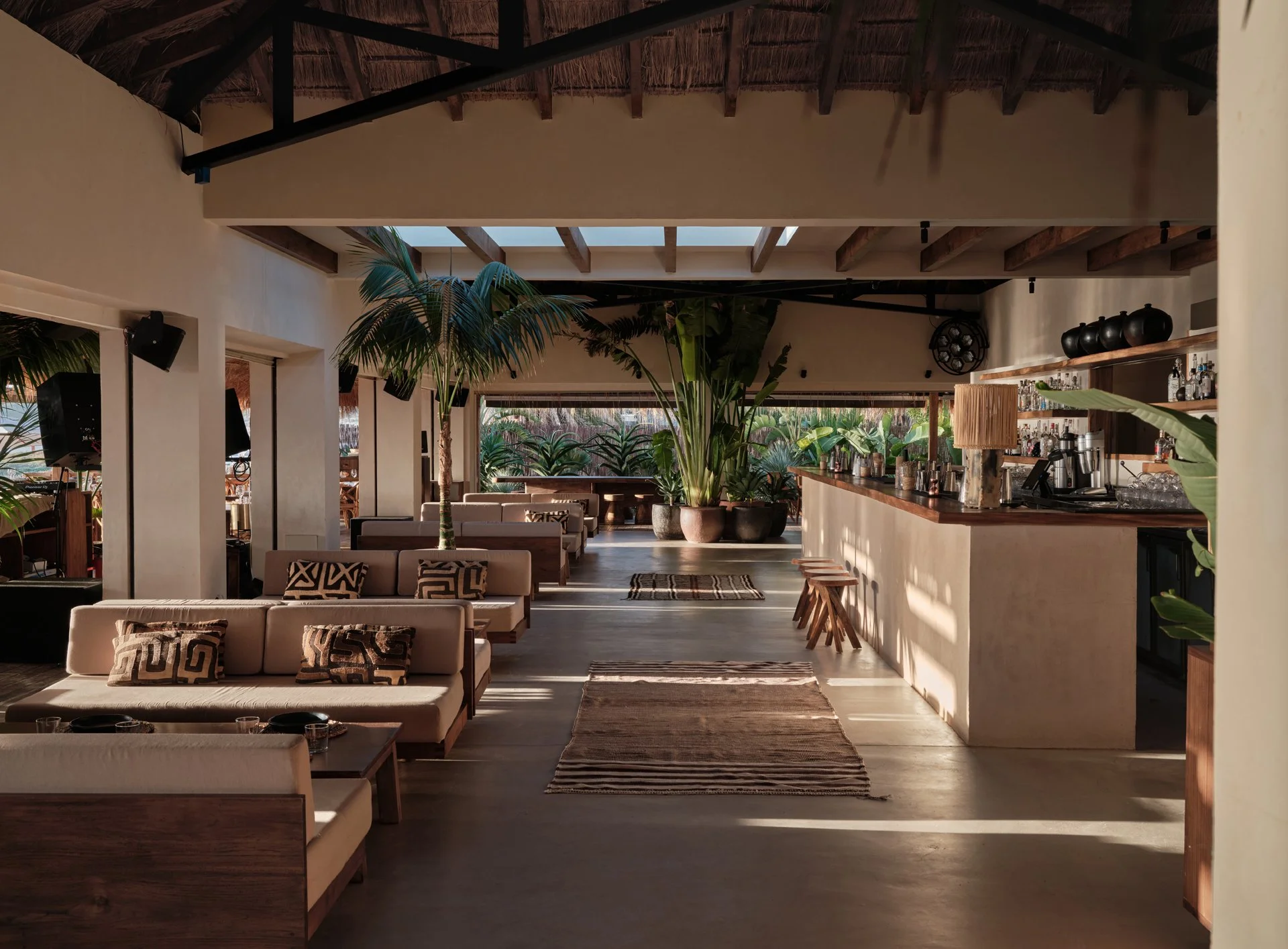 Stylish beach club interior with lounge seating, wooden bar, and tropical plants under a high, open ceiling with natural light.