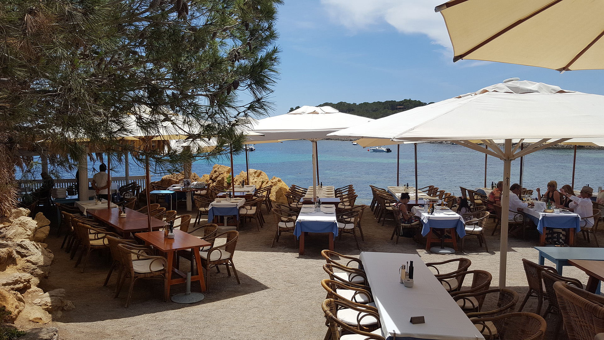 Seaside restaurant terrace with tables and chairs under large parasols, overlooking calm blue water and rocky coastline.