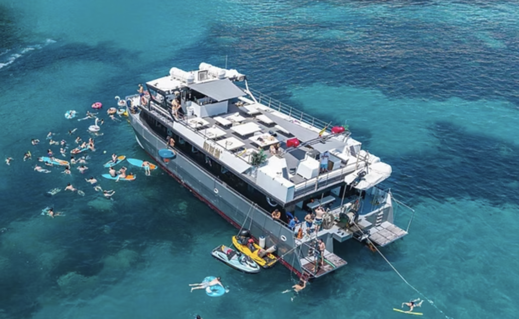 Luftaufnahme eines großen Bootes, das in klarem, türkisfarbenem Wasser vor Anker liegt, während Menschen an einem sonnigen Tag schwimmen, auf aufblasbaren Gegenständen treiben und sich rund um das Boot entspannen.