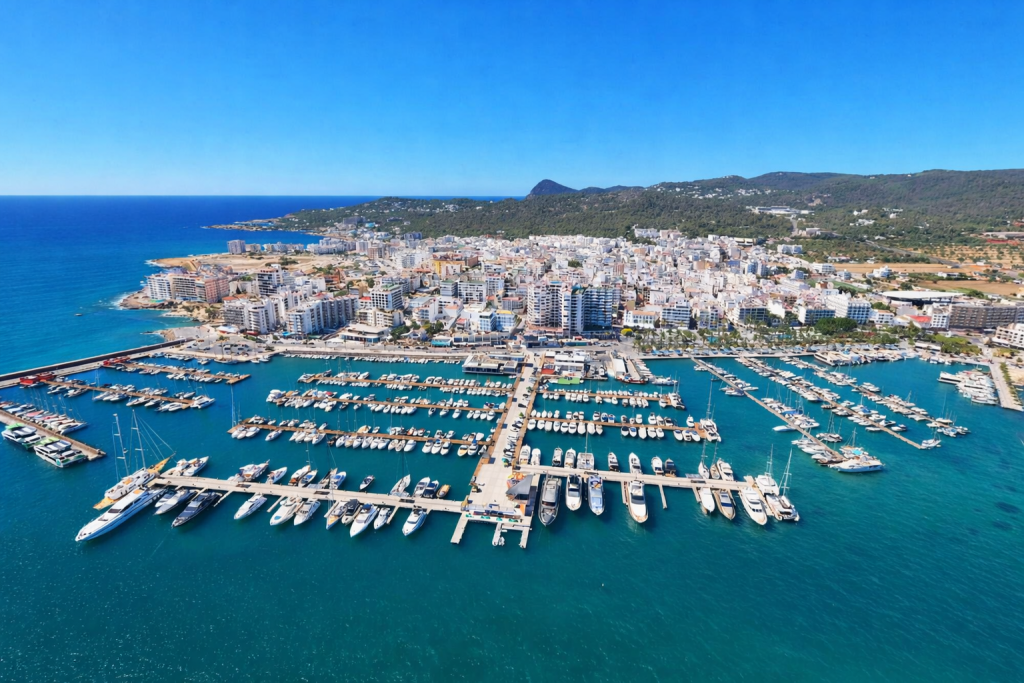 Aerial view of a coastal Mediterranean town with a large marina filled with yachts and boats, turquoise water, dense white buildings along the shoreline, and green hills in the background under a clear blue sky.