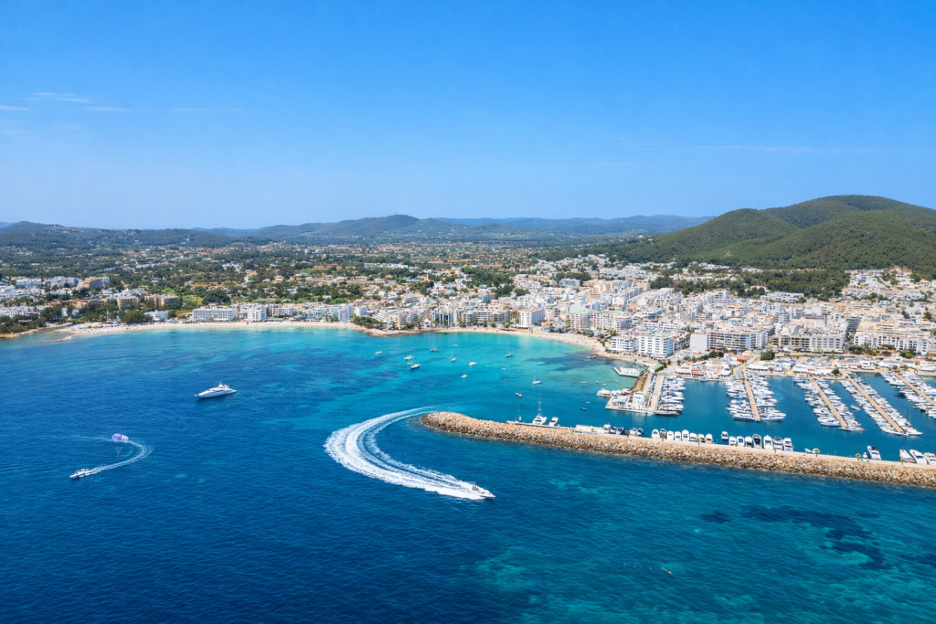 Aerial view of a Mediterranean coastline with a curved bay, turquoise water, a marina filled with boats, a speedboat leaving a white trail, and a coastal town with white buildings set against green hills under a clear blue sky.
