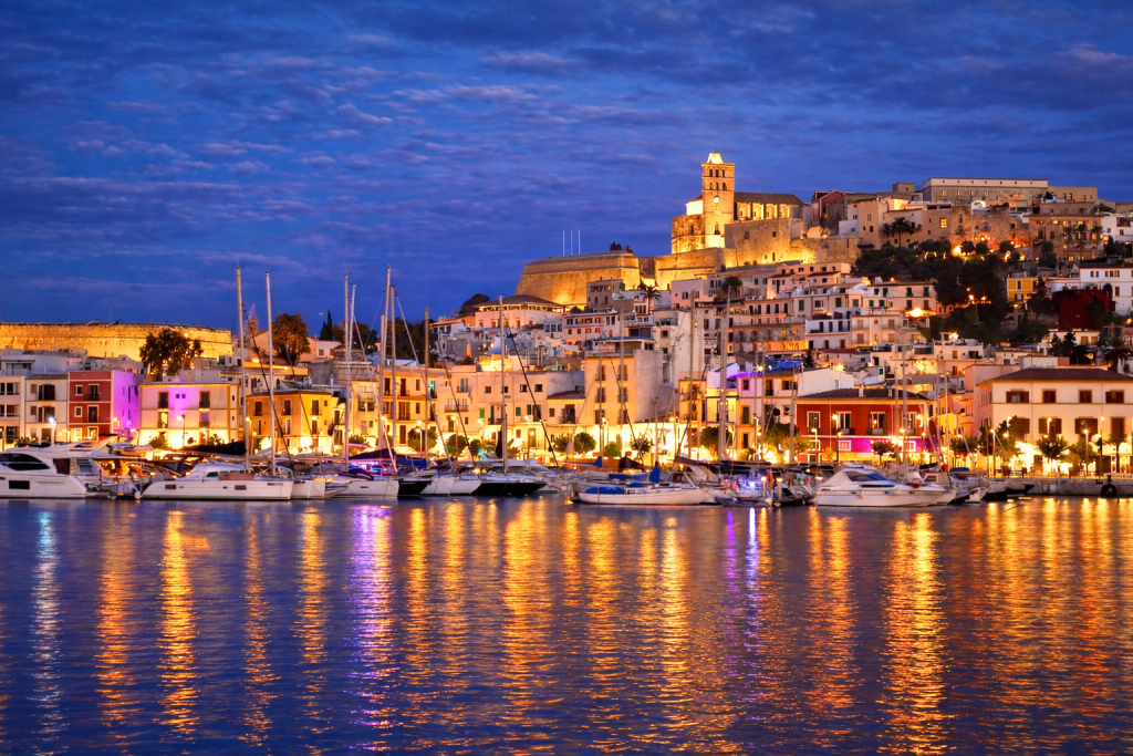 Twilight view of a Mediterranean harbour town with white hillside buildings and a lit historic fortress, yachts and sailboats docked along the marina, and colourful reflections shimmering across calm water.