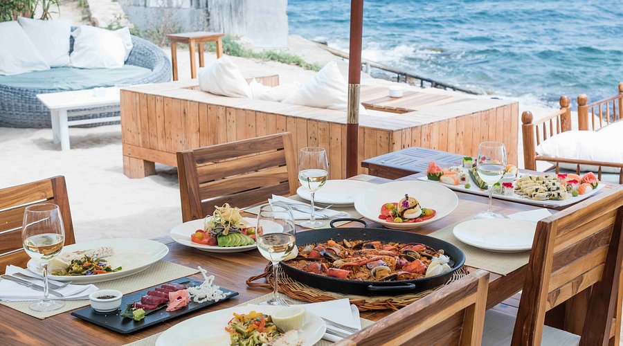 Seafront dining table with fresh seafood and dishes, set beside the water with wooden seating and relaxed beach lounge area in the background.