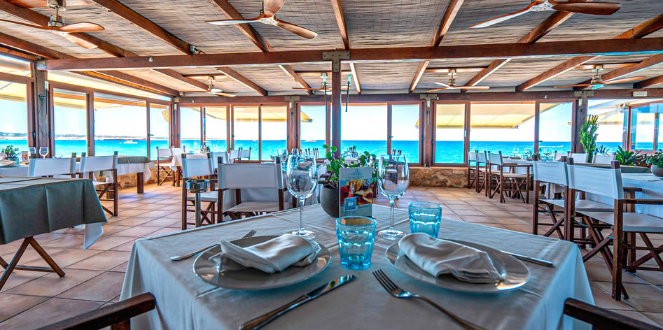 Seafront restaurant interior with wooden beams and large windows, set tables with white tablecloths, overlooking the ocean.
