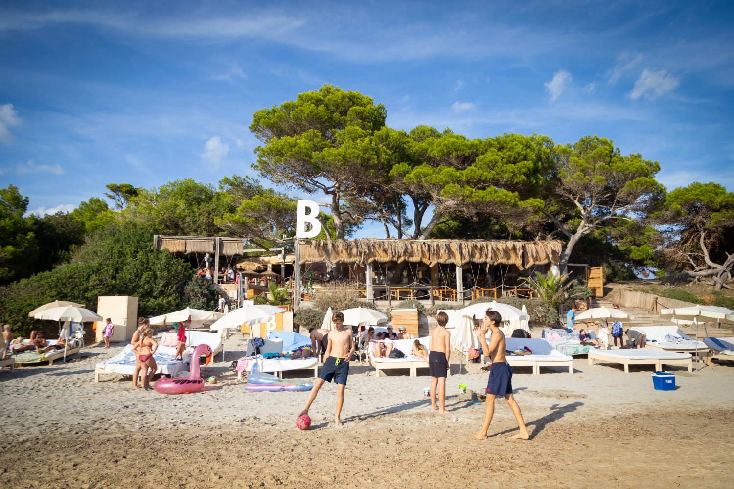 Busy beach club with sunbeds and parasols, people relaxing and children playing on the sand in front of a rustic, tree-lined beachfront restaurant.