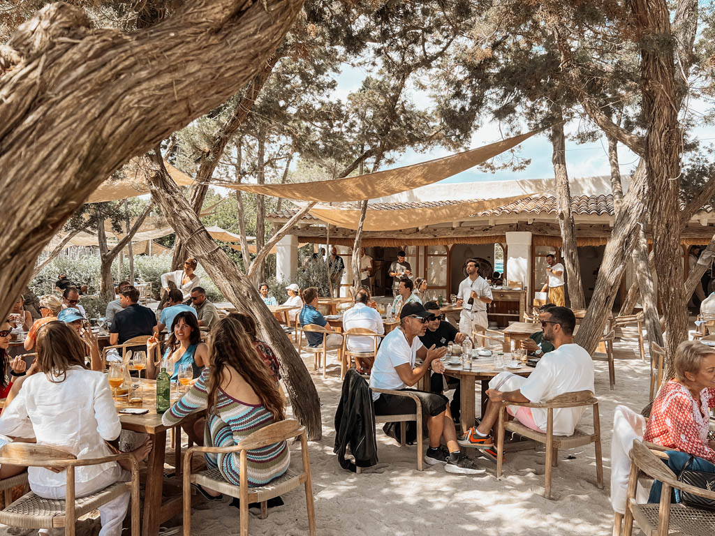 Outdoor beach restaurant set among trees with shaded seating, wooden tables, and people dining in a relaxed, sandy setting.
