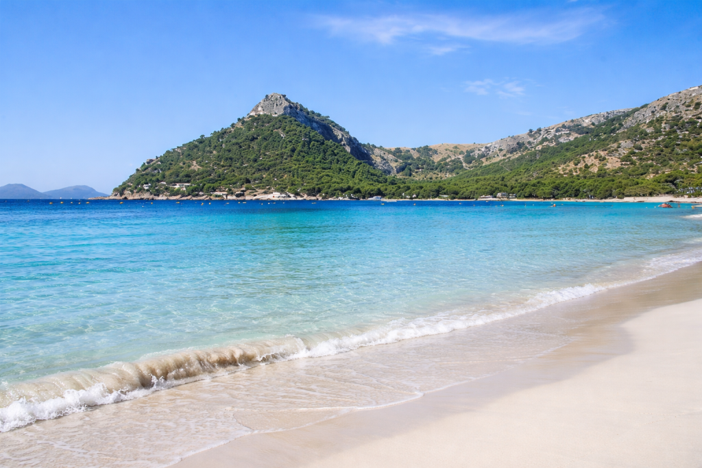 Mer turquoise limpide léchant une plage de sable fin aux vagues douces, adossée à une montagne verdoyante et à une côte rocheuse sous un ciel d'un bleu éclatant.