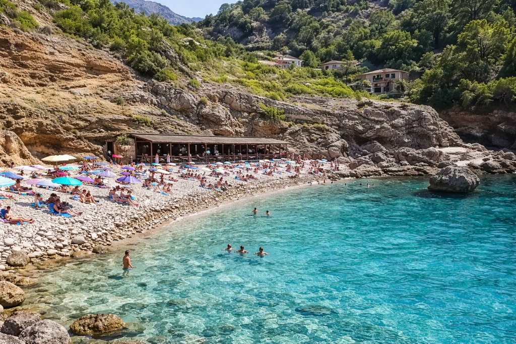 Crique turquoise aux eaux claires, nageurs près du rivage, plage de galets animée bordée de falaises rocheuses, bar de plage rustique et villas à flanc de colline entourées de pins sous un ciel d'un bleu éclatant.
