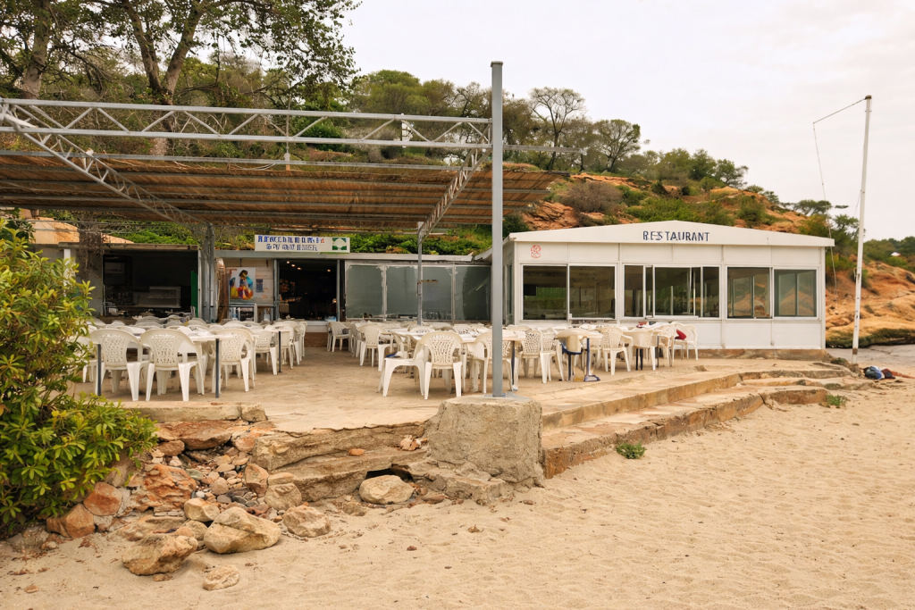 Casual beachside restaurant with white plastic chairs and shaded outdoor seating, set on sandy ground beside a simple building and coastal hillside.