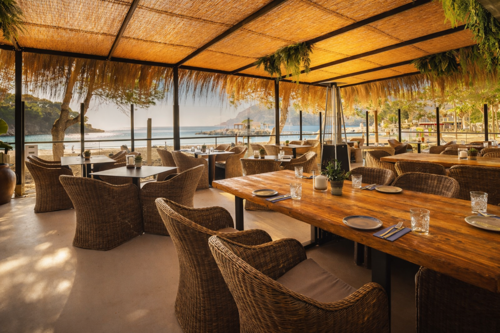 Seaside restaurant terrace with wicker chairs and wooden tables under a bamboo canopy, overlooking a calm beach and sea in warm evening light.