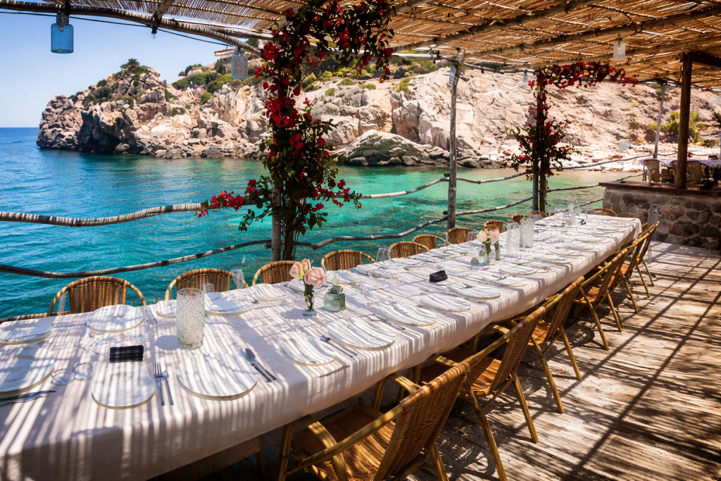 Seaside dining terrace with a long table set under a shaded pergola, overlooking clear turquoise water and rocky cliffs, with flowers wrapped around wooden beams.