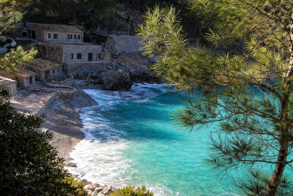 Secluded Mediterranean cove with turquoise water, small pebble beach and rustic stone houses nestled among pine trees, with waves gently reaching the shore