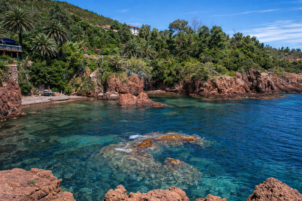 Rocky coastal cove with clear blue water and red stone formations, surrounded by lush greenery and hillside homes overlooking the shoreline.