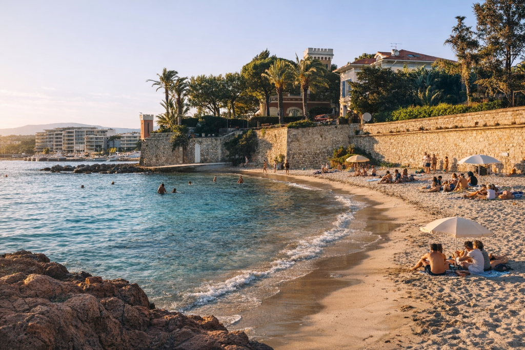 Sunny seaside beach with people relaxing and swimming, backed by a stone wall and palm-lined villas overlooking the coastline.