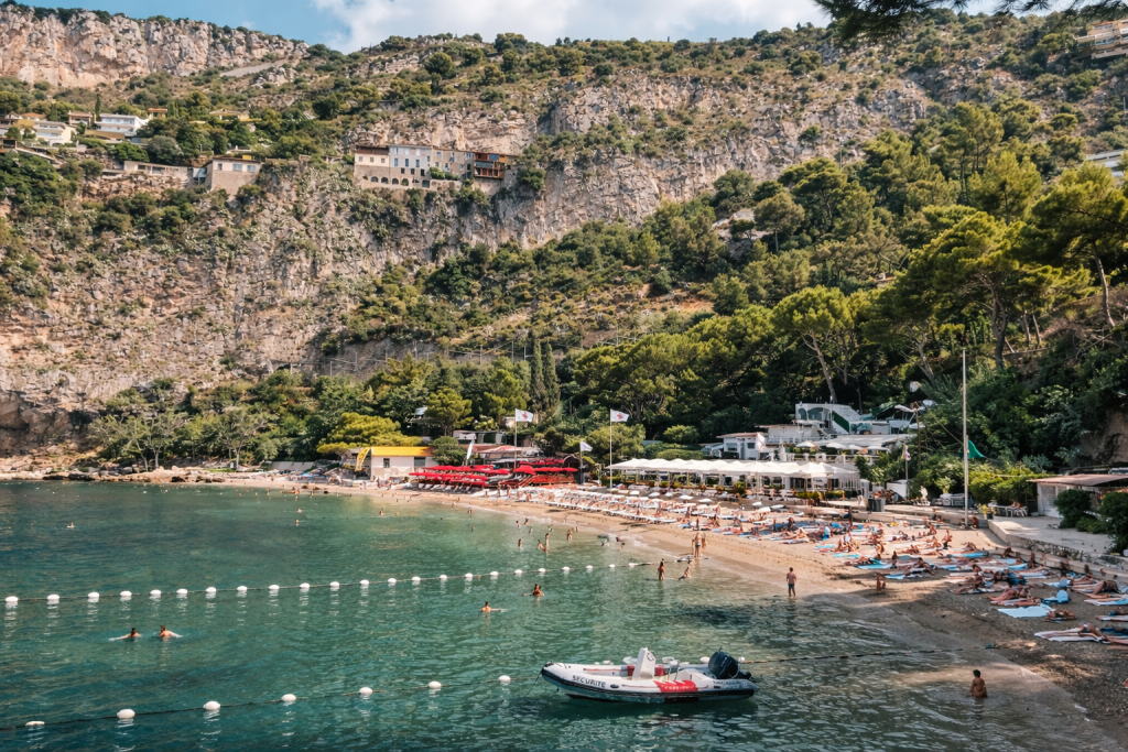 Mediterranean beach cove with swimmers and sunbathers, lined with loungers and umbrellas, set beneath steep cliffs with villas and lush greenery above.