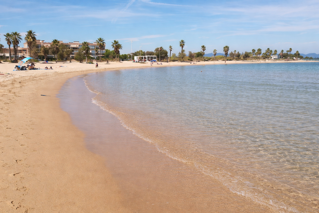 Wide sandy beach with calm shallow water, lined with palm trees and low buildings, under a clear blue sky with a few people relaxing along the shore.
