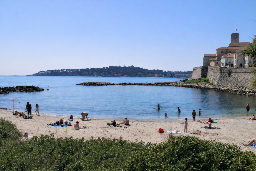 Sandy beach with people relaxing and swimming in calm water, bordered by stone walls and historic buildings, with a distant coastline across the bay.