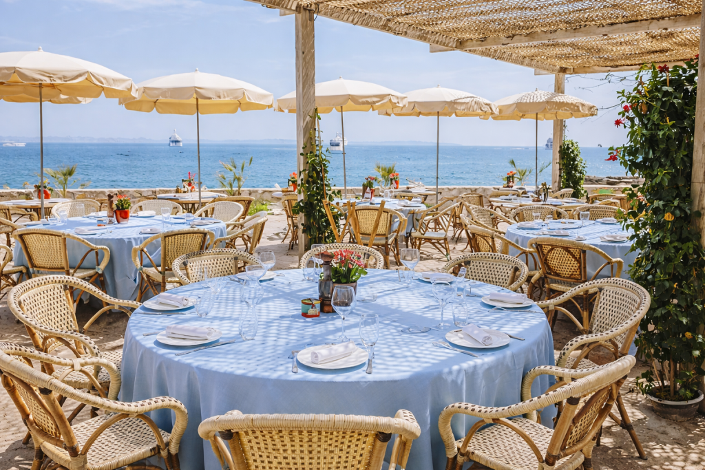 Terraza de un restaurante junto al mar con mesas redondas y manteles azules, sillas de mimbre y grandes sombrillas color crema, con vistas al océano y barcos en la distancia.