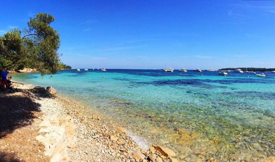 Rocky shoreline with clear turquoise water and anchored boats in the distance, framed by trees and a bright blue sky along a calm coastal bay.